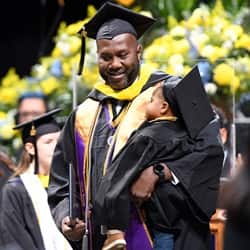 A father carrying his child at the Fall 2022 Commencement. Both are dressed in graduation caps and gowns.