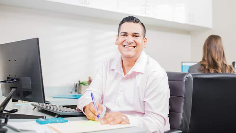 Juan Munoz, who earned his degree from SNHU in 2022, sitting in an office next to a computer, writing in a  notebook with a woman with brown hair facing away from him in the background.