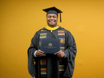 Aaron Davenport, who earned his online master's in finance from SNHU in 2025, wearing his cap and gown and holding his diploma in front of a yellow backdrop.