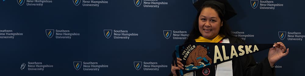 SNHU graduate and Alaska resident Stella Bory in her cap and gown holding an Alaska pennant in front of an SNHU step and repeat backdrop.