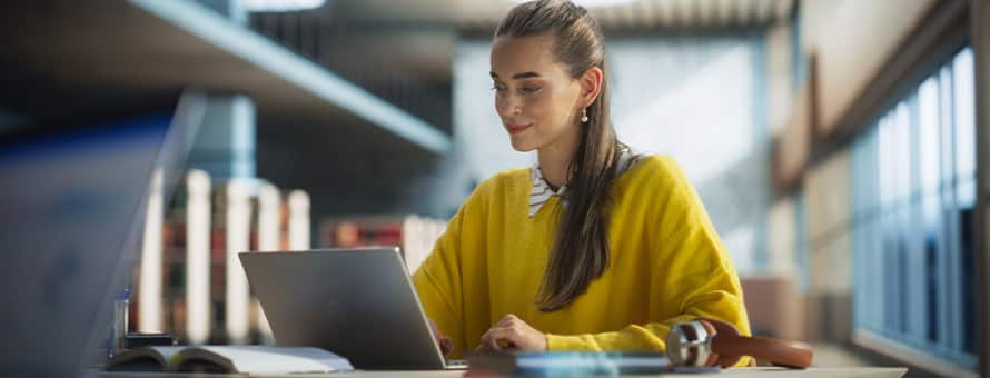 A student wearing a yellow sweater, using a laptop in a public library to earn her master's degree online.