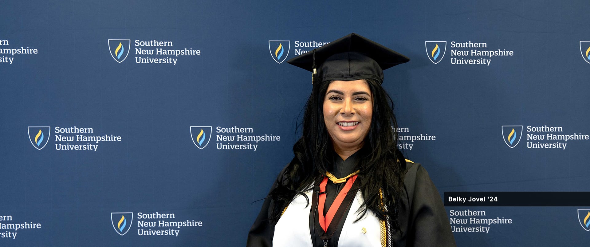 Belky Jovel, who earned her associate's in accounting from SNHU in 2024, wearing her cap and gown standing in front of a backdrop featuring SNHU's logo.