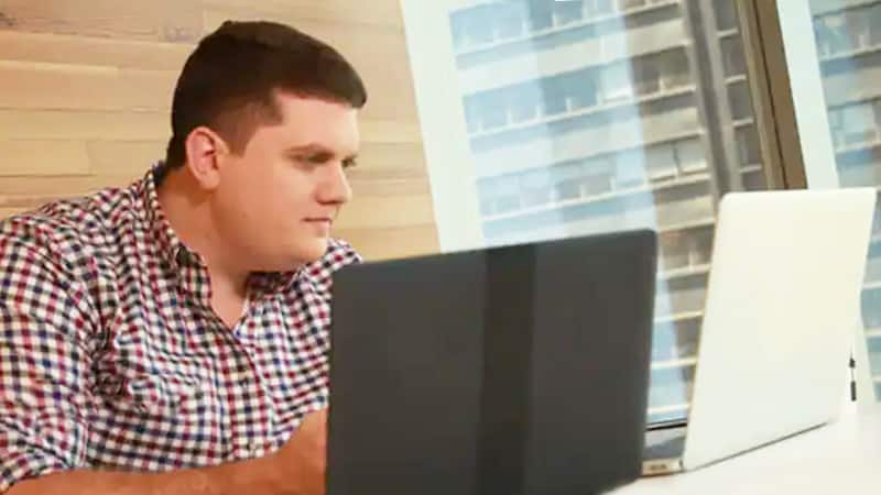 Kyle Turner, who earned his degree in 2015, sitting in front of a large window with two laptops open in front of him.