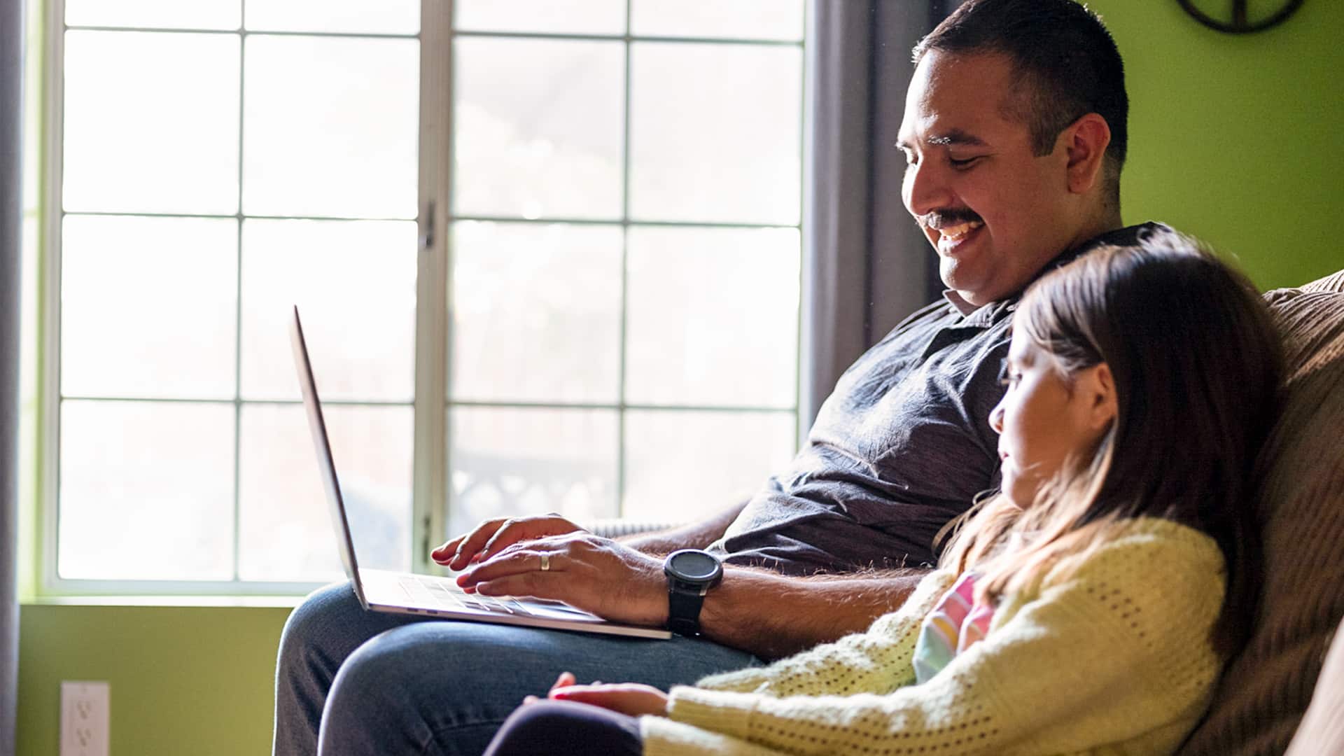 Salvador Villa, who earned his AS in Criminal Justice in 2019, sitting on a couch smiling and working on his laptop in front a large bright window and with a young girl sitting next to him.