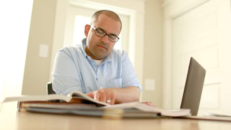 Alex Romero, who earned his degree from SNHU in 2017, wearing a button-down shirt working on his laptop and reading an open textbook on the table next to him. 