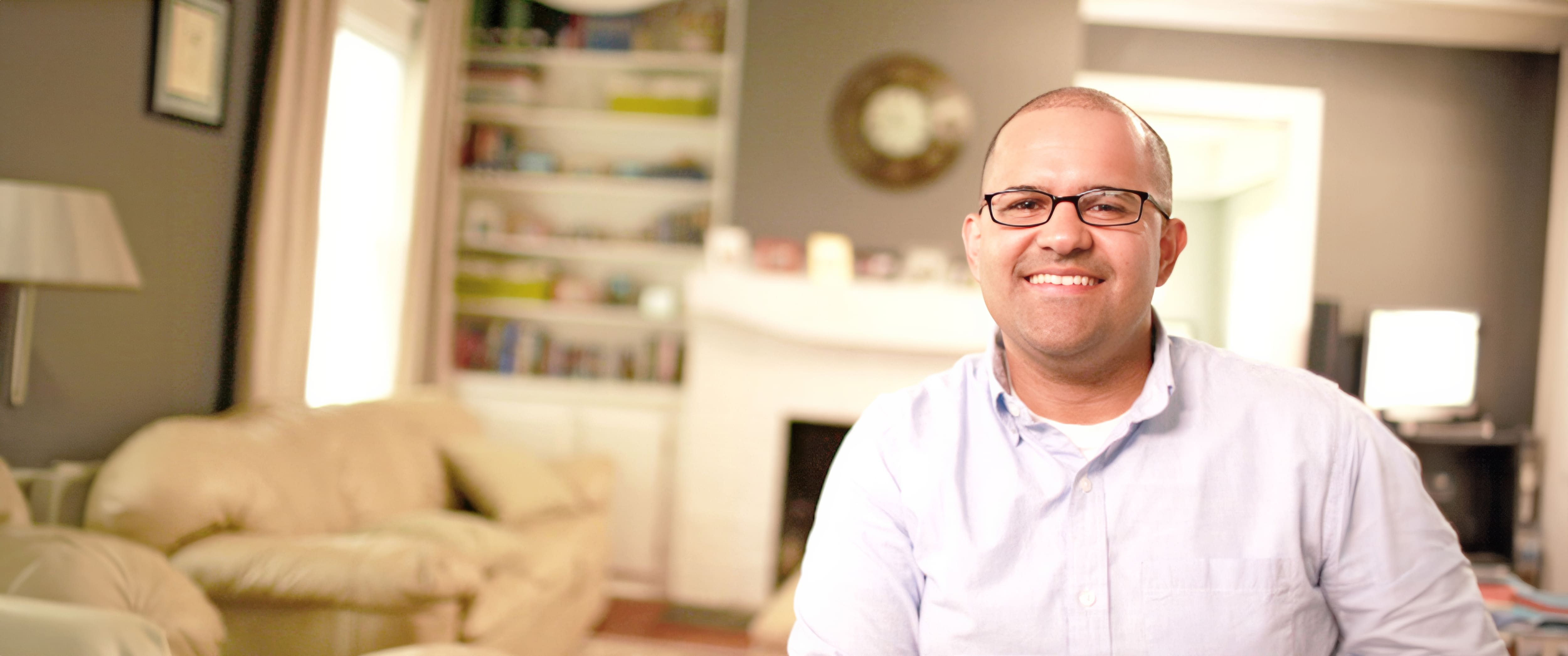 Alex Romero, who earned his degree from SNHU in 2017, wearing a buttondown shirt and eyeglasses in his living room with a white leather couch in the background.