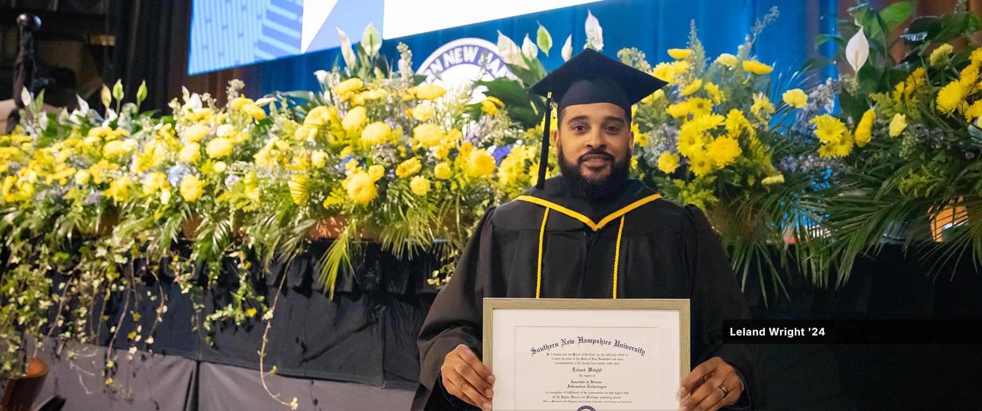 Leland Wrigher, who earned his associate in IT from SNHU in 2024, wearing his cap and gown, holding his framed diploma and standing in front of a row of bright yellow flowers.
