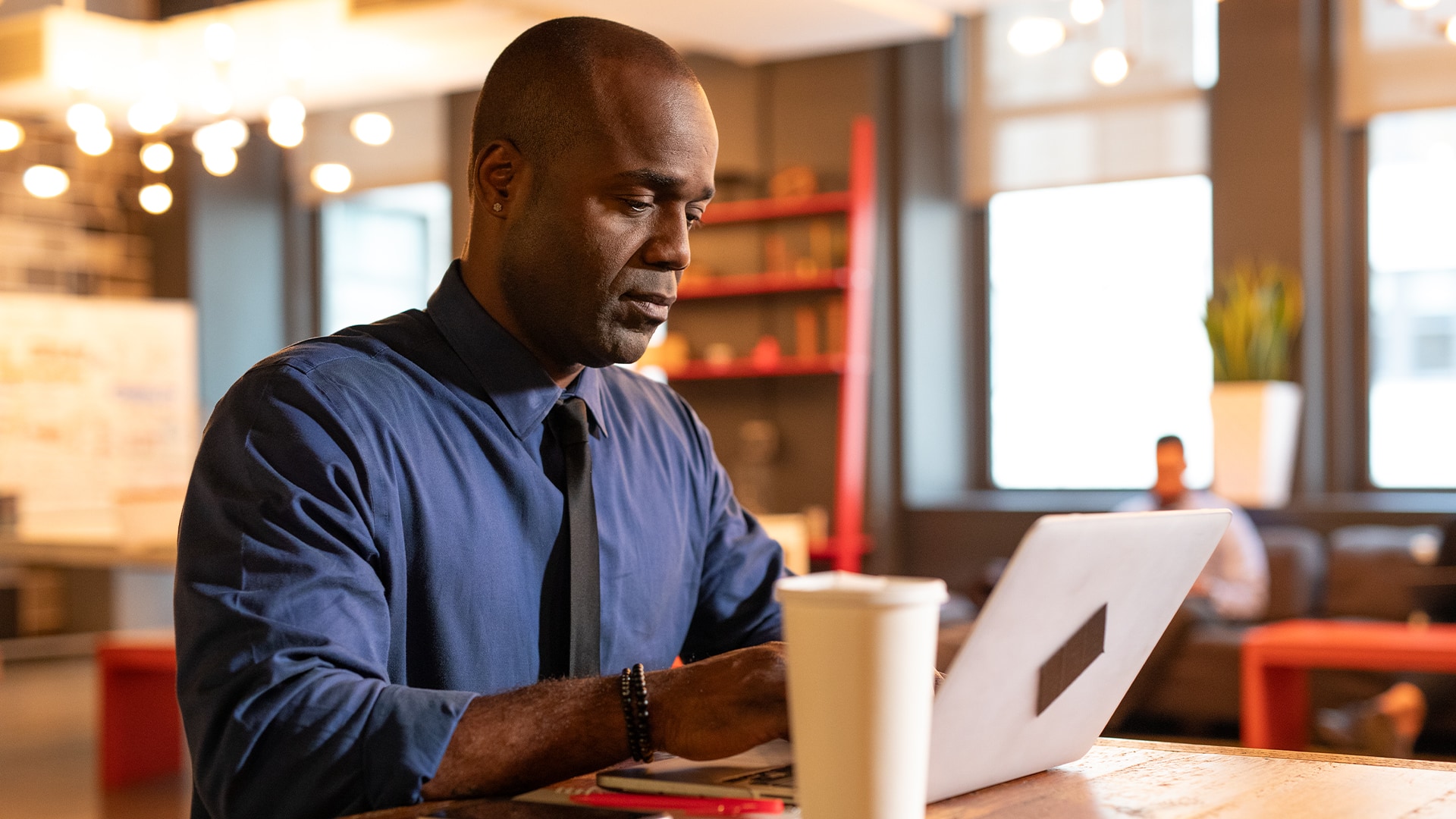 Jean Besson, who earned a degree from SNHU in 2014, wearing a blue buttondown shirt and black tie working on his  laptop in a coffee shop with a cup of coffee on the table beside him