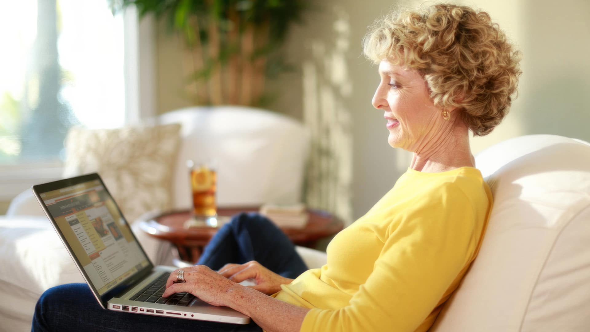 Susan Ryan, who earned her degree from SNHU, wearing a bright yellow shirt, sitting on a counch and working on her  laptop with a glass of ice tea on a small table beside her.
