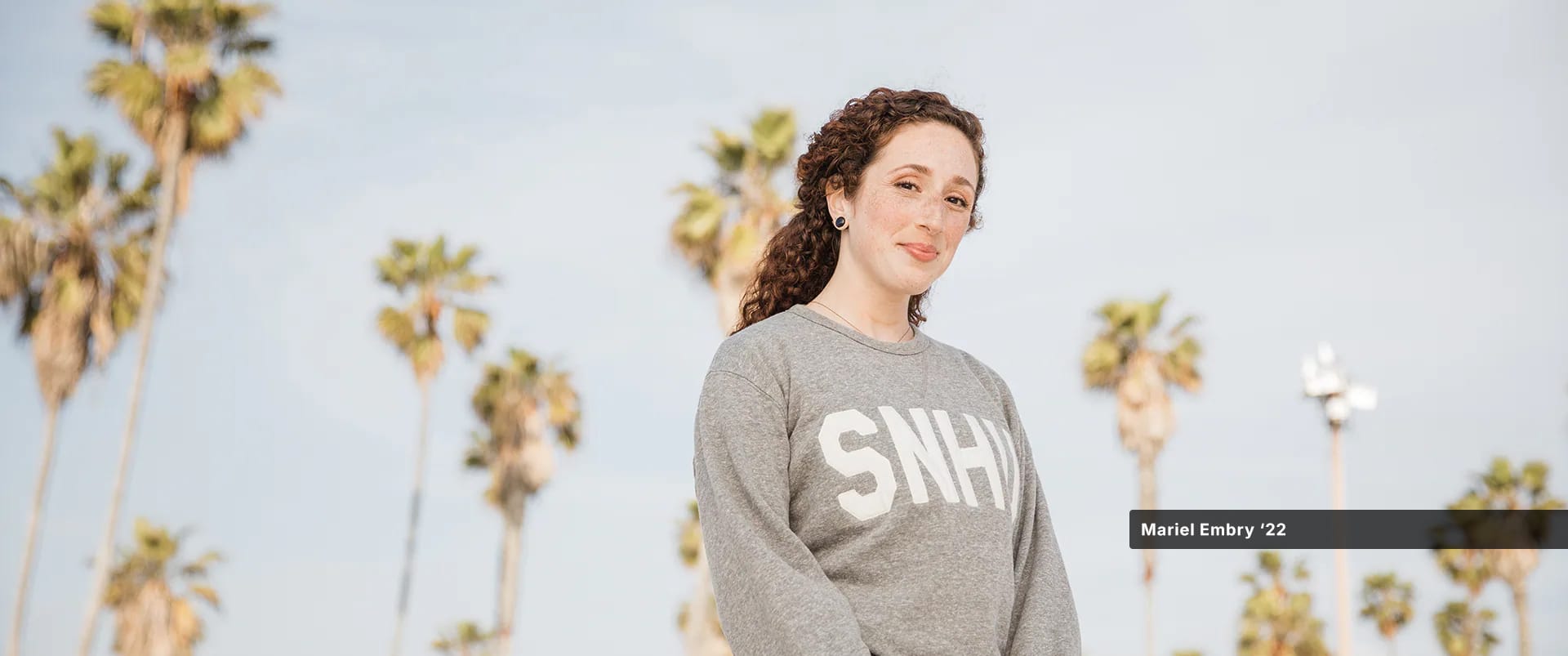 Mariel Embry, who earned her degree in creative writing and English from SNHU, wearing a grey SNHU sweatshirt with tall palm trees behind her.
