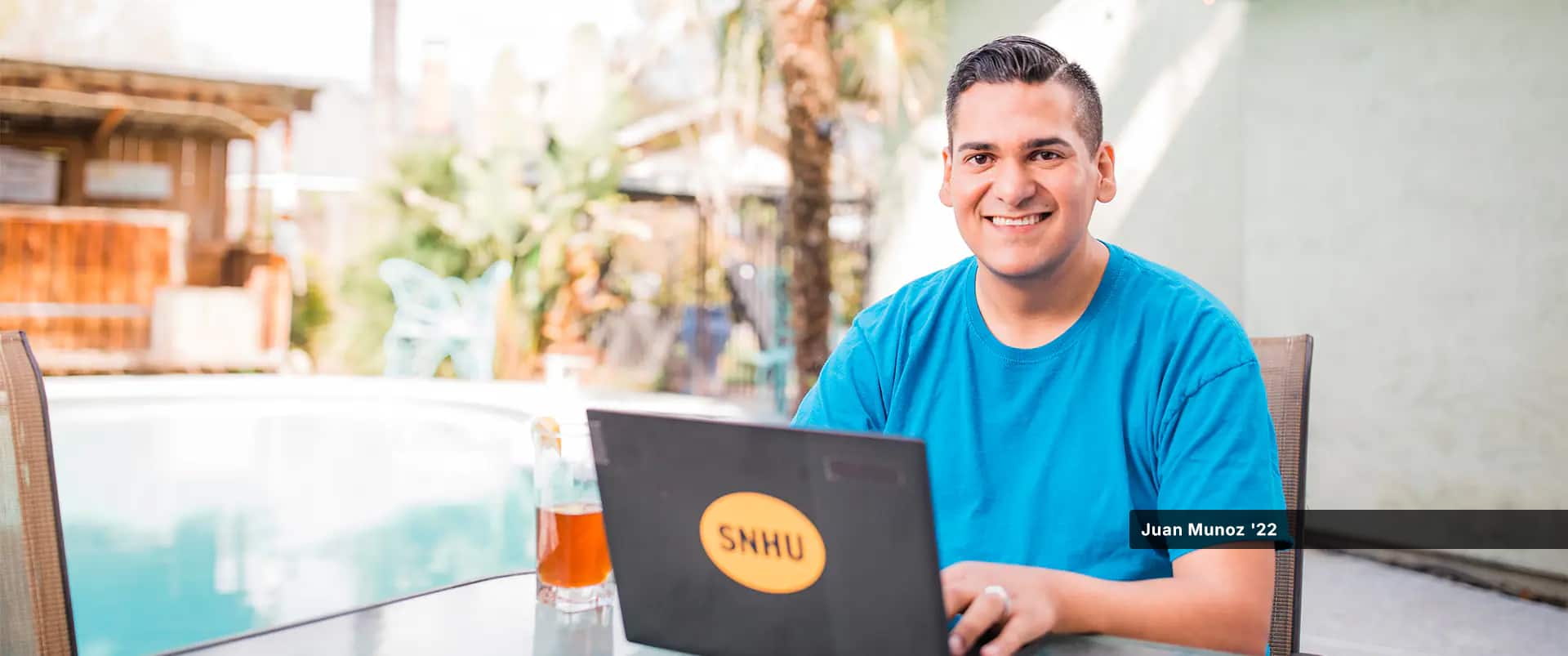 Juan Munoz, who earned his general studies degree in 2022, smiling and working on his laptop beside a pool and with a glass of ice tea with a lemon beside him.