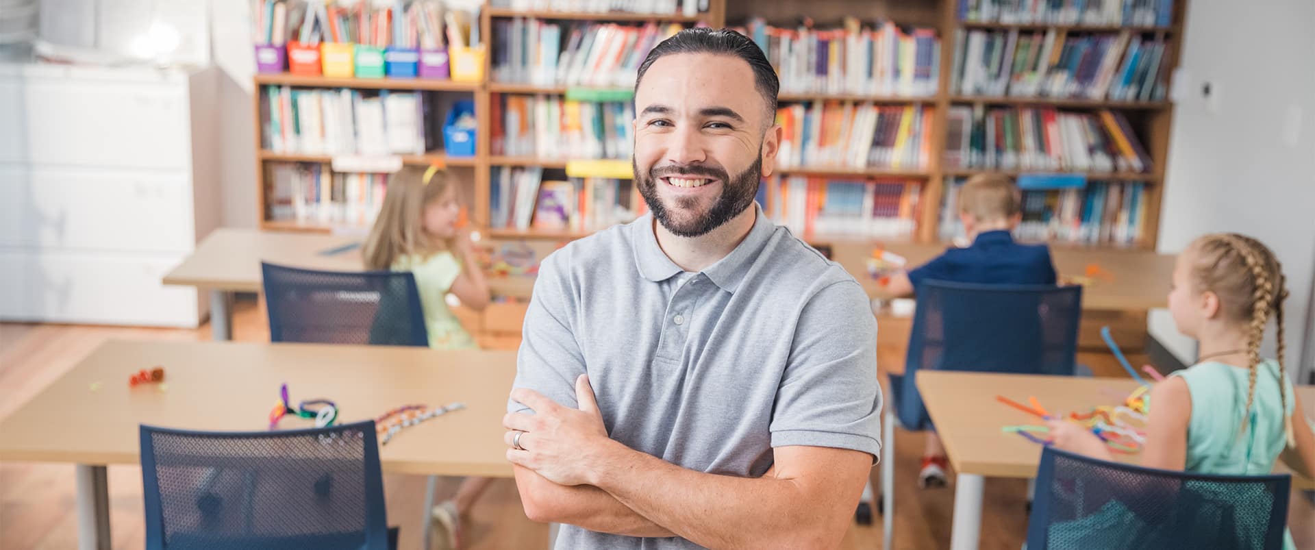 Stephen Goulakos, a 2022 psychology degree online graduate, standing in the middle of a classroom with young children playing at their desks.