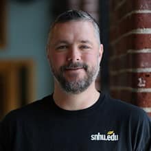 Ben Steinbach, a career outreach specialist at SNHU, wearing a dark SNHU T-shirt in front of a brick column.