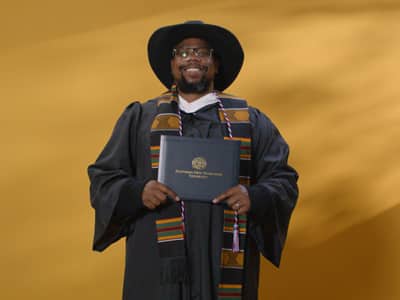 Brandon Hayes, who earned his bachelor's in graphic design from SNHU in 2024, wearing a cowboy hat and cap and gown and holding his diploma.