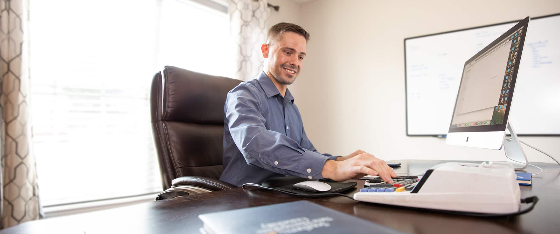 Blake Venable with a computer keyboard and calculator on a desk in front of him and a whiteboard on the wall in the background.