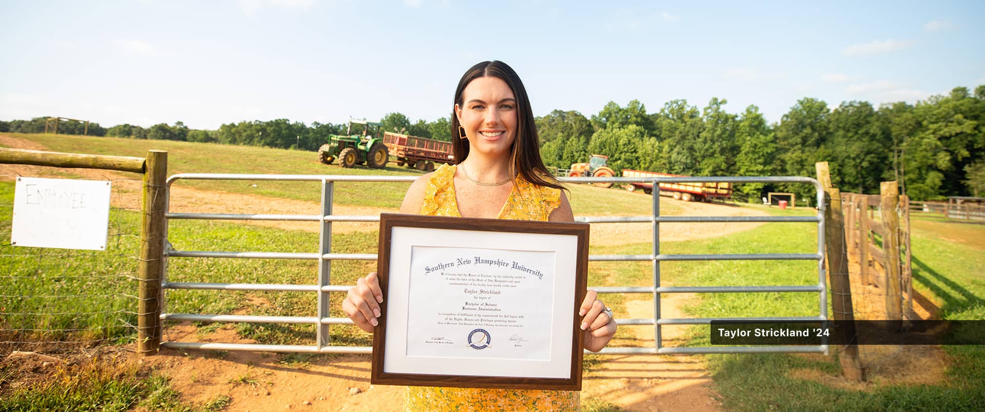 Taylor Strickland, who earned her bachelor's in business administration from SNHU in 2024, holding her framed diploma and standing in front of a wood and metal fence with two tractors in a field behind her.