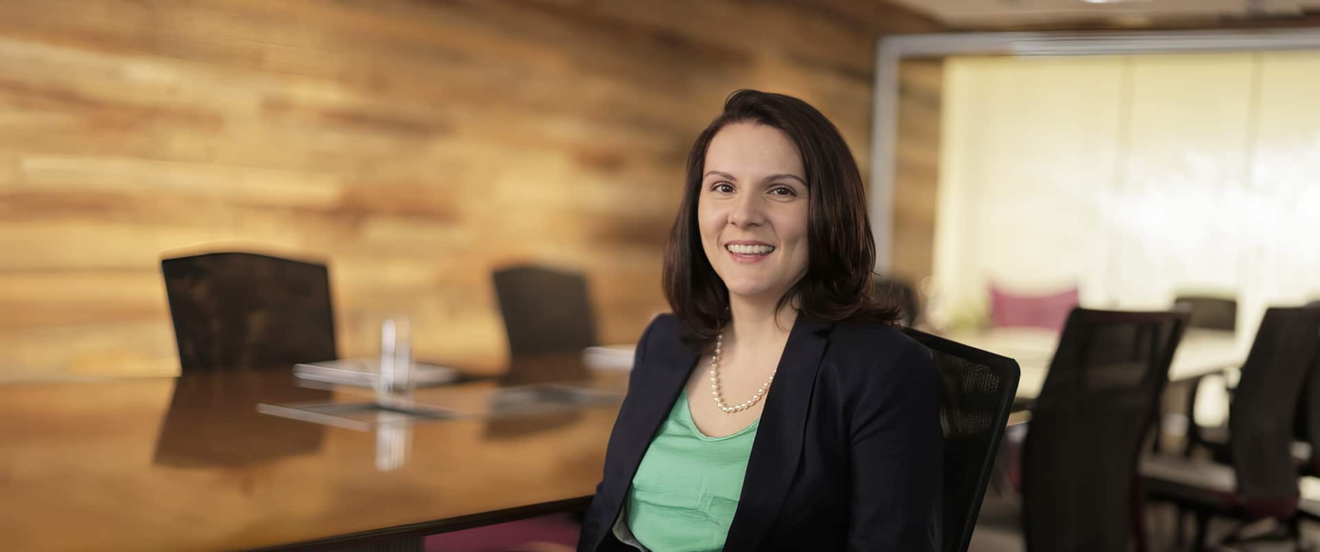 Jessye Giley wearing a light green blouse, dark blazer and pearl necklace sitting in a wood  paneled conference room with a large table and several chairs in the background.