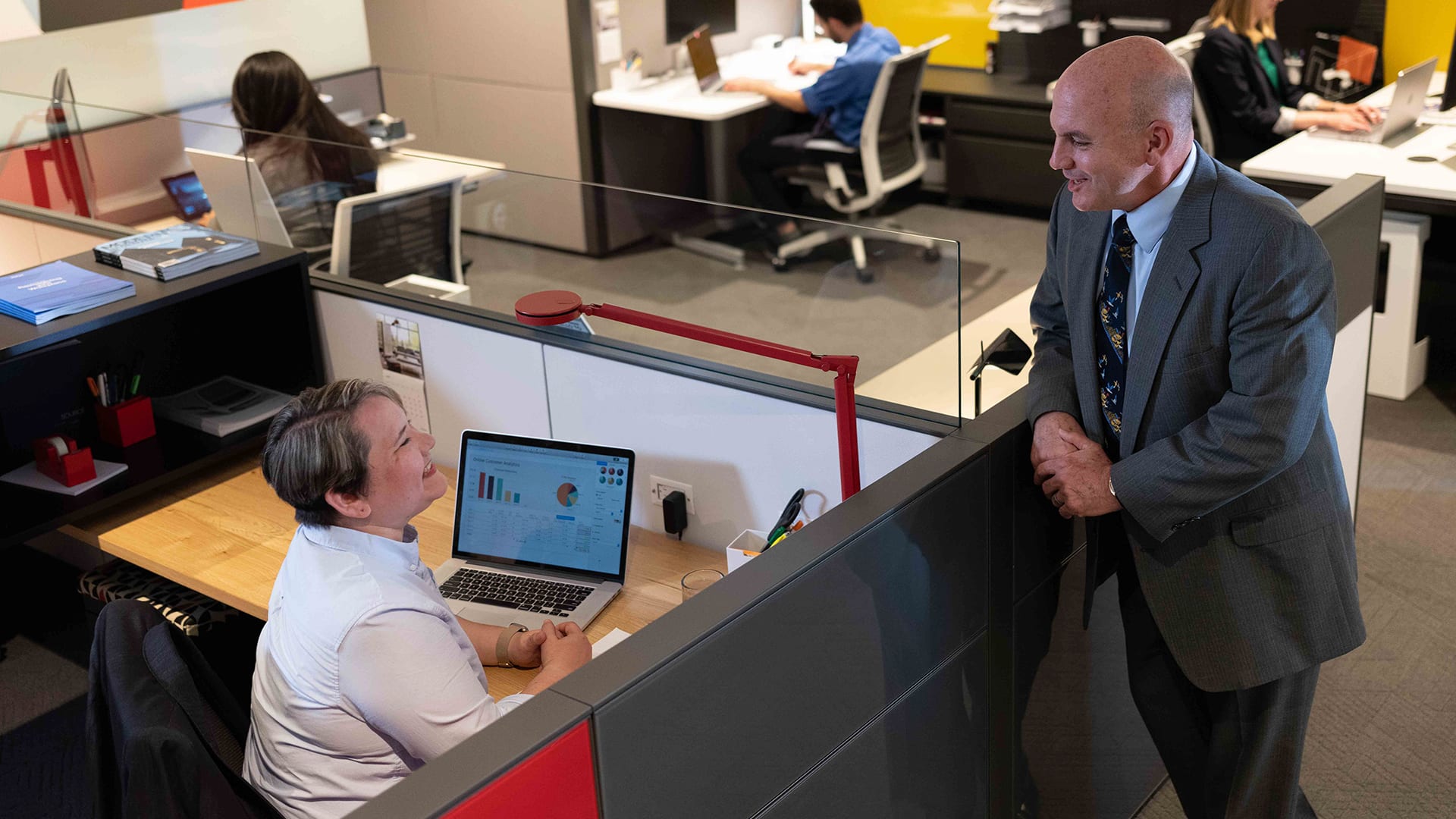 Alaine Garcia, who earned her degree in 2020, in an office cubicle with an open laptop showing charts and graphs while she talks to a man standing nearby.