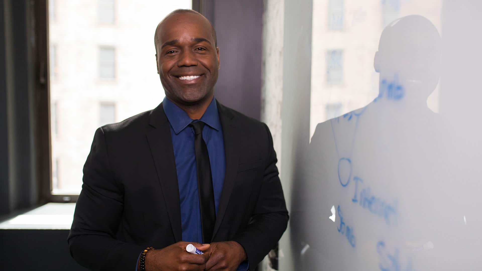 Jean Besson, who earned his degree from SNHU in 2014, wearing a dark suit and tie, holding a blue dry-erase marker  and standing next to a white board.