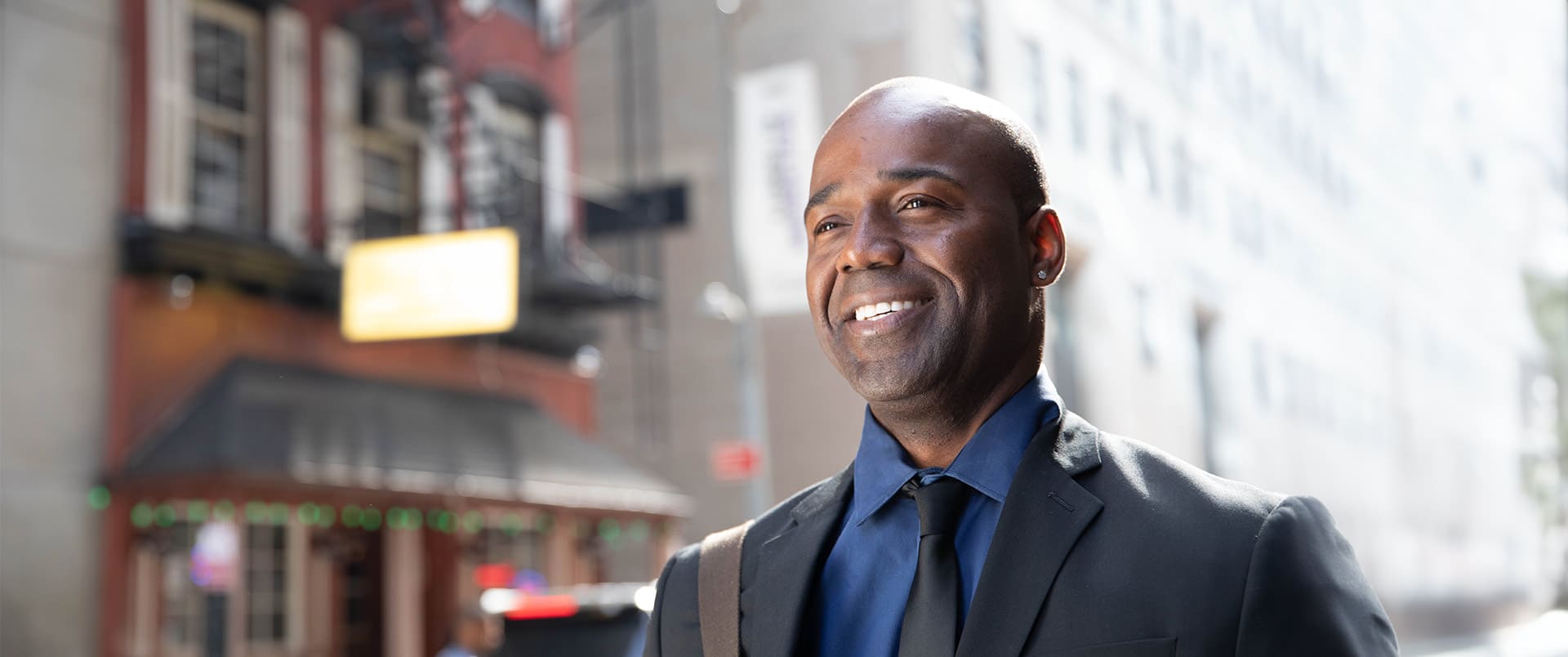Jean Besson, who earned his degree from SNHU in 2014, wearing a suit and tie walking  down a city street with a shoulder bag on his right shoulder.