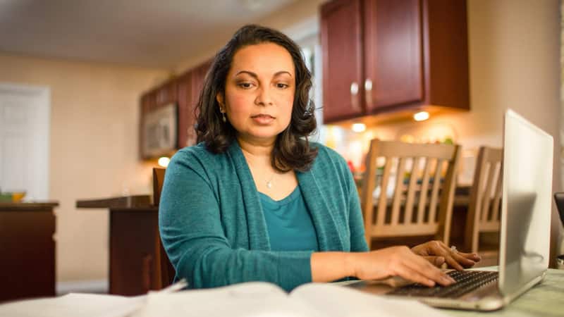 Katrina Gomes, who earned her degree from SNHU, wearing a green blouse and sweater sitting at her kitchen table working on her laptop.