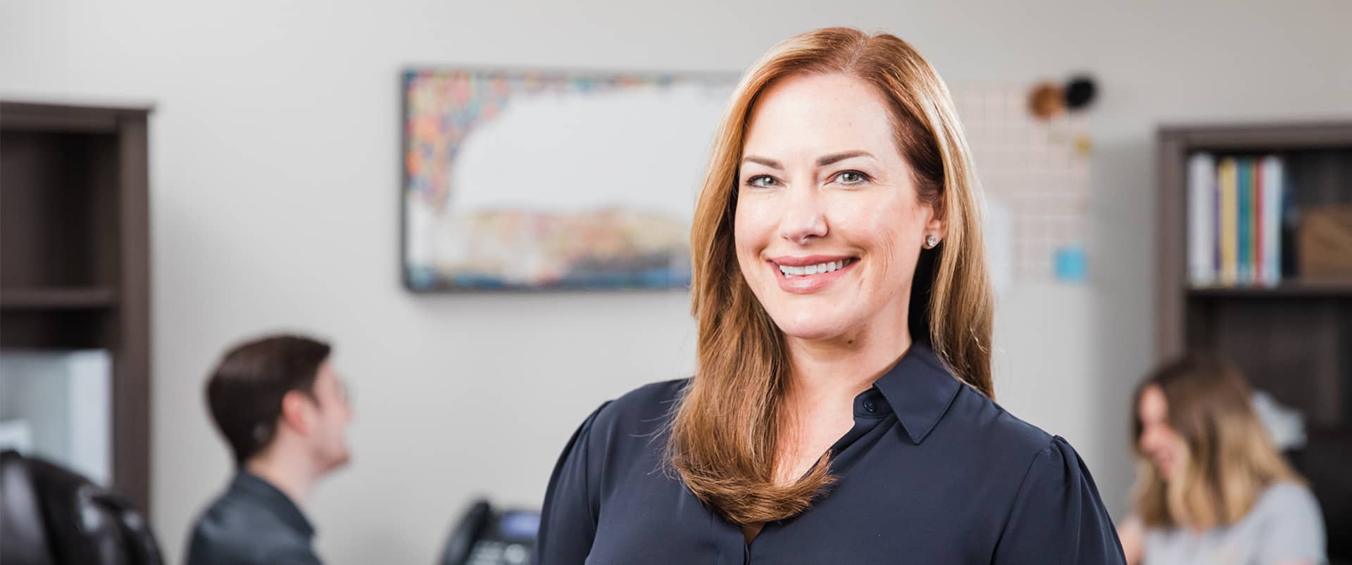 Nikki Bennet, who earned her degree from SNHU, wearing a dark blouse in an office with two people and a bookcase in the background.