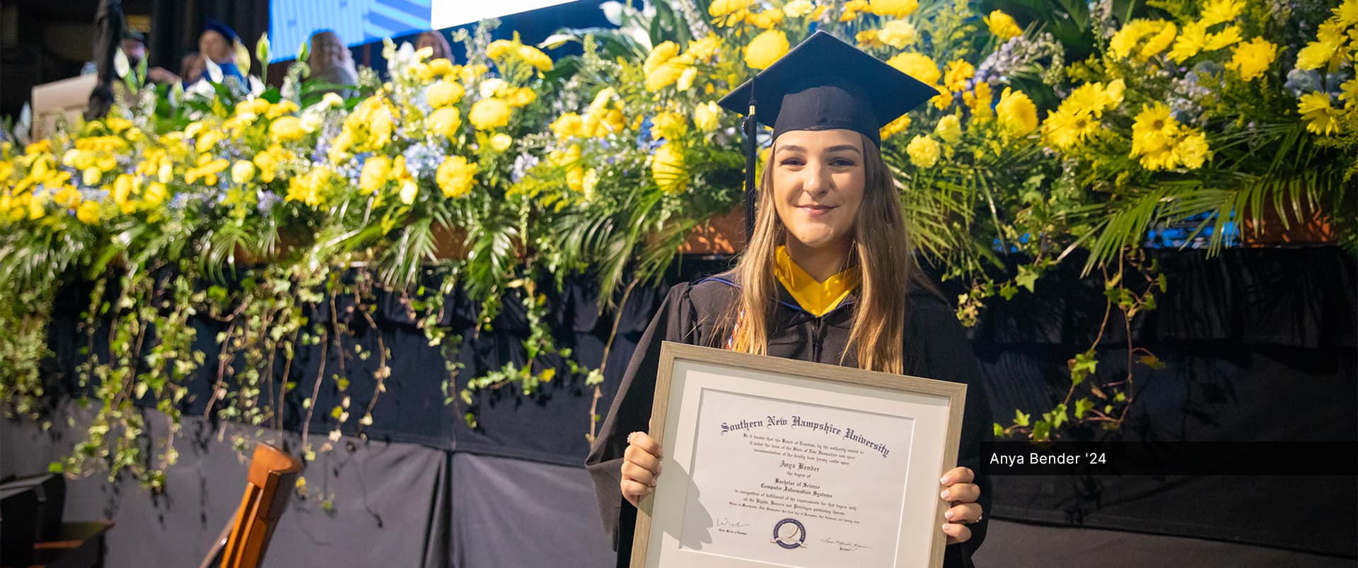 Anya Bender, who earned her bachelor's in computer information systems from SNHU in 2024, wearing her cap and gown and holding her framed diploma with a row of bright yellow flowers behind her.