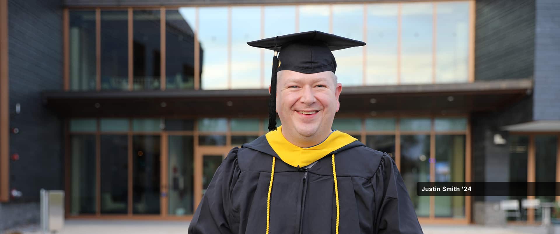 Justin Smith, who earned his bachelor's in cybersecurity from SNHU in 2024, wearing his cap and gown standing in front of a campus building.
