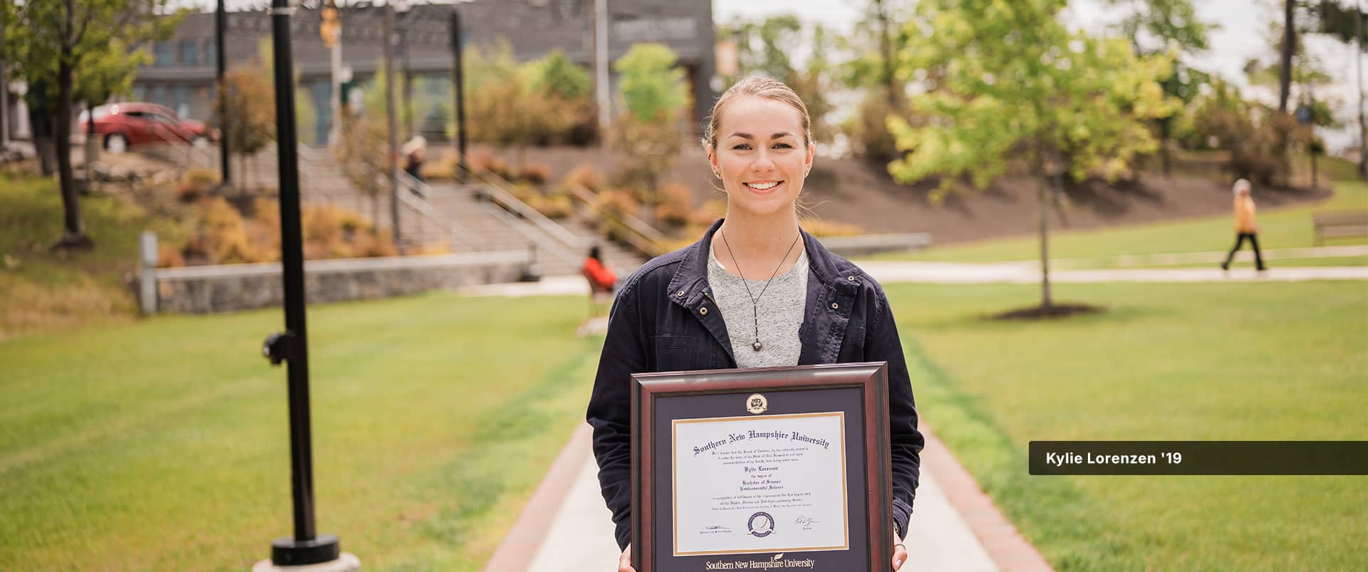 Kylie Lorenzen, who earned her degree from SNHU in 2019, holding her framed diploma while standing in the quad on the SNHU campus.