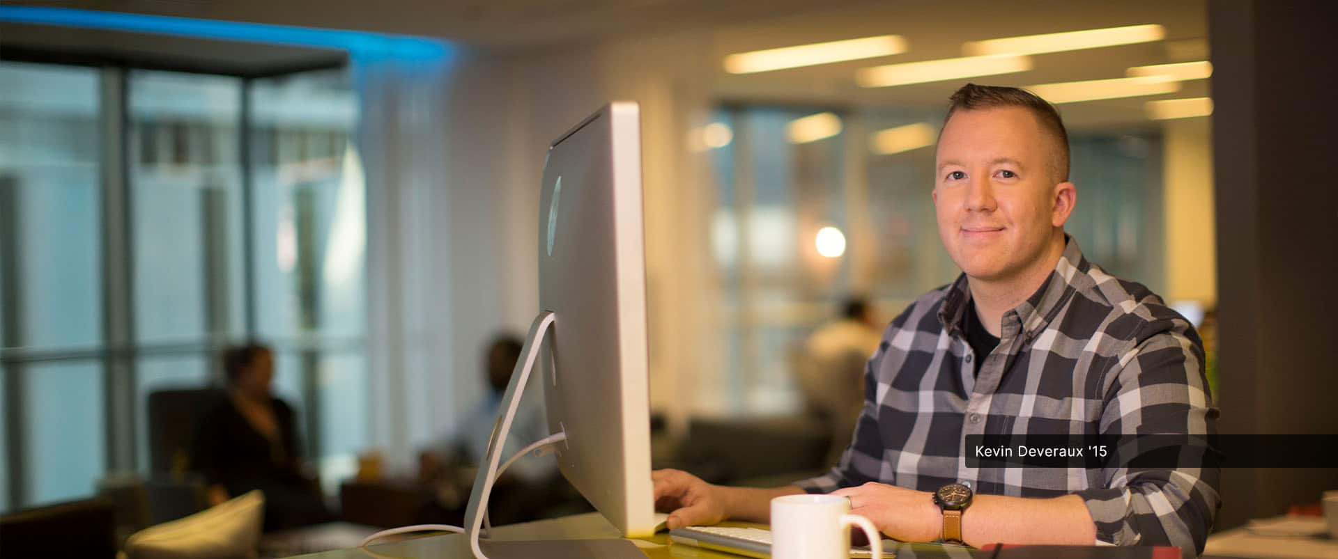 Kevin Deveraux, an SNHU alum, wearing a black and grey plaid button down shirt at a computer with two thin notebooks, a smartphone and white coffee mug next to him.