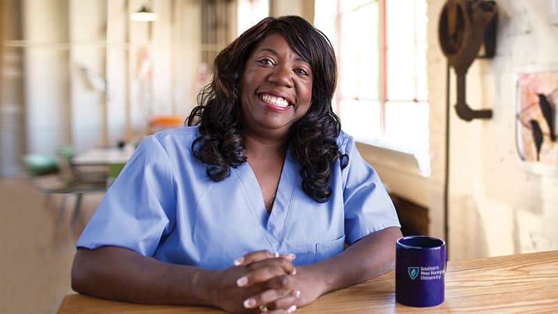 Dana Aulds, who earned her degree from SNHU in 2015, sitting at a table, wearing light blue scrubs with a blue Southern New Hampshire University coffee mug to her left.