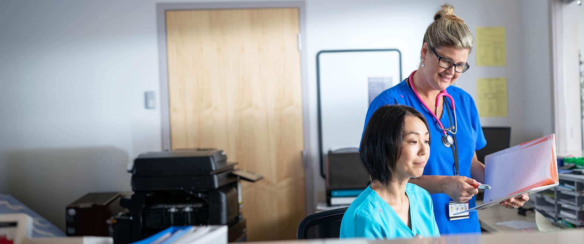 Kristina Libby, who earned her degree from SNHU in 2017, wearing blue nurse scrubs and a pink stethoscope around her neck standing next to a seated woman also wearing scrubs.
