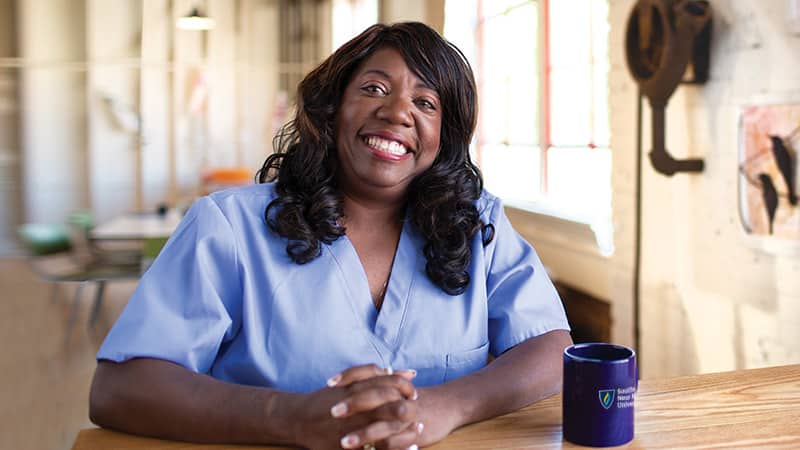 Dana Aulds, an online degree graduate from SNHU, sitting in scrubs at a desk in a healthcare office.