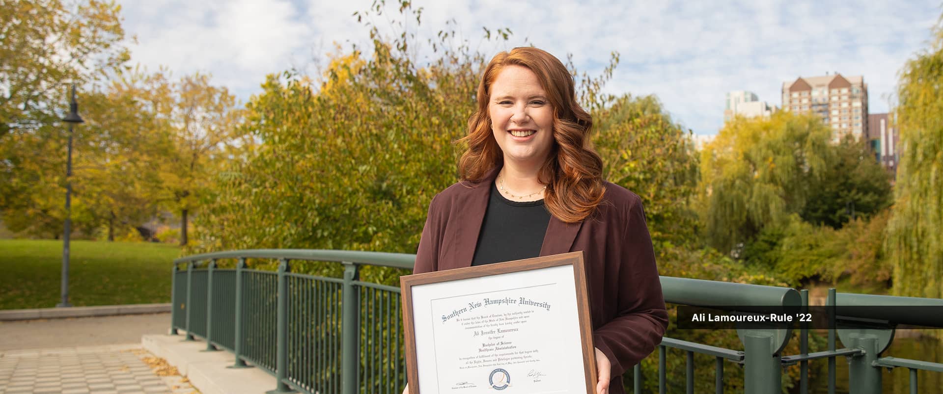 Ali Lamoureux-Rule holding her 2022 BS in Healthcare Administration diploma while standing on a footbridge in a park.