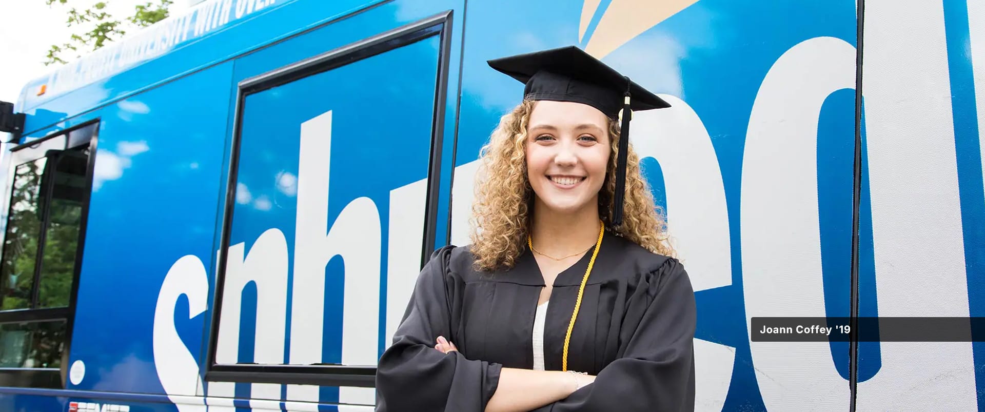 Joann Coffey, who earned her degree from SNHU, wearing her cap and gown and standing in front of the SNHU-branded bus.