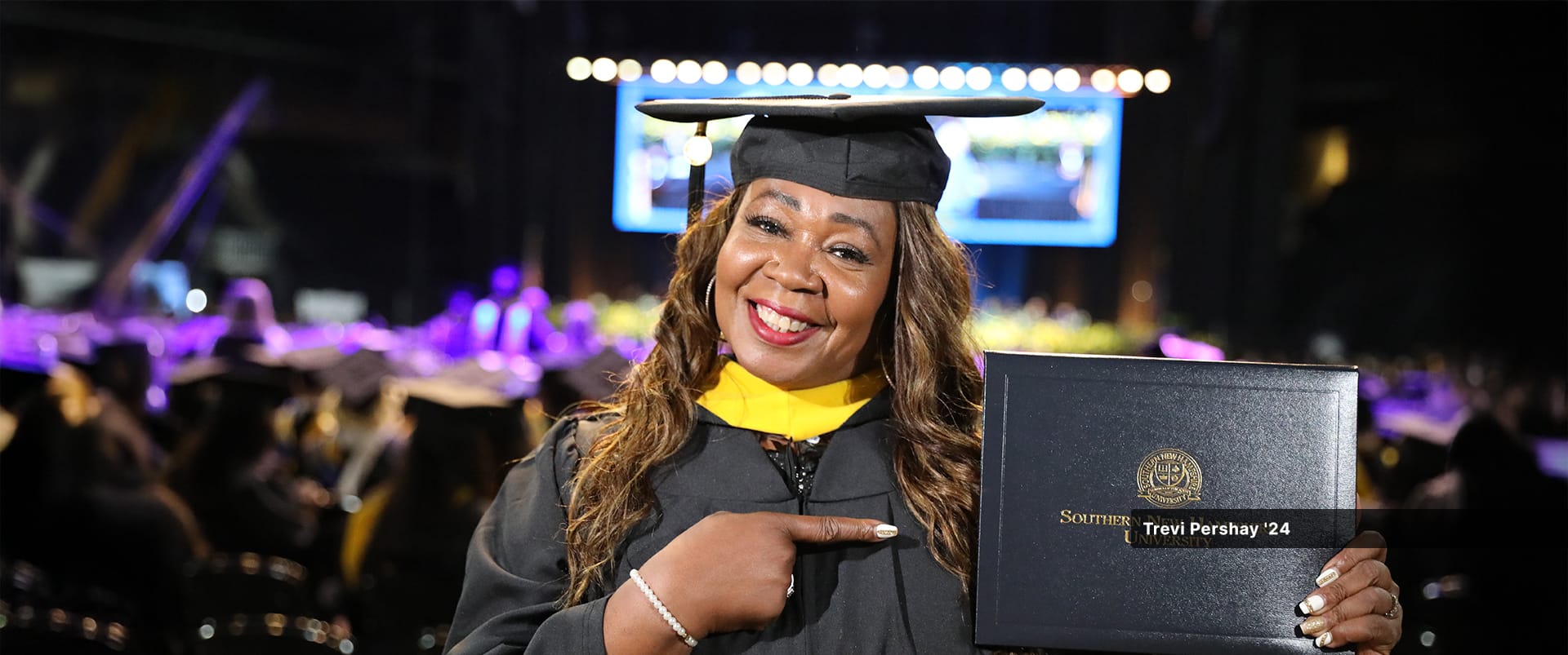 Trevi Pershay, who earned her bachelor's in marketing from SNHU in 2024, wearing her cap and gown, holding up and pointing at her diploma.