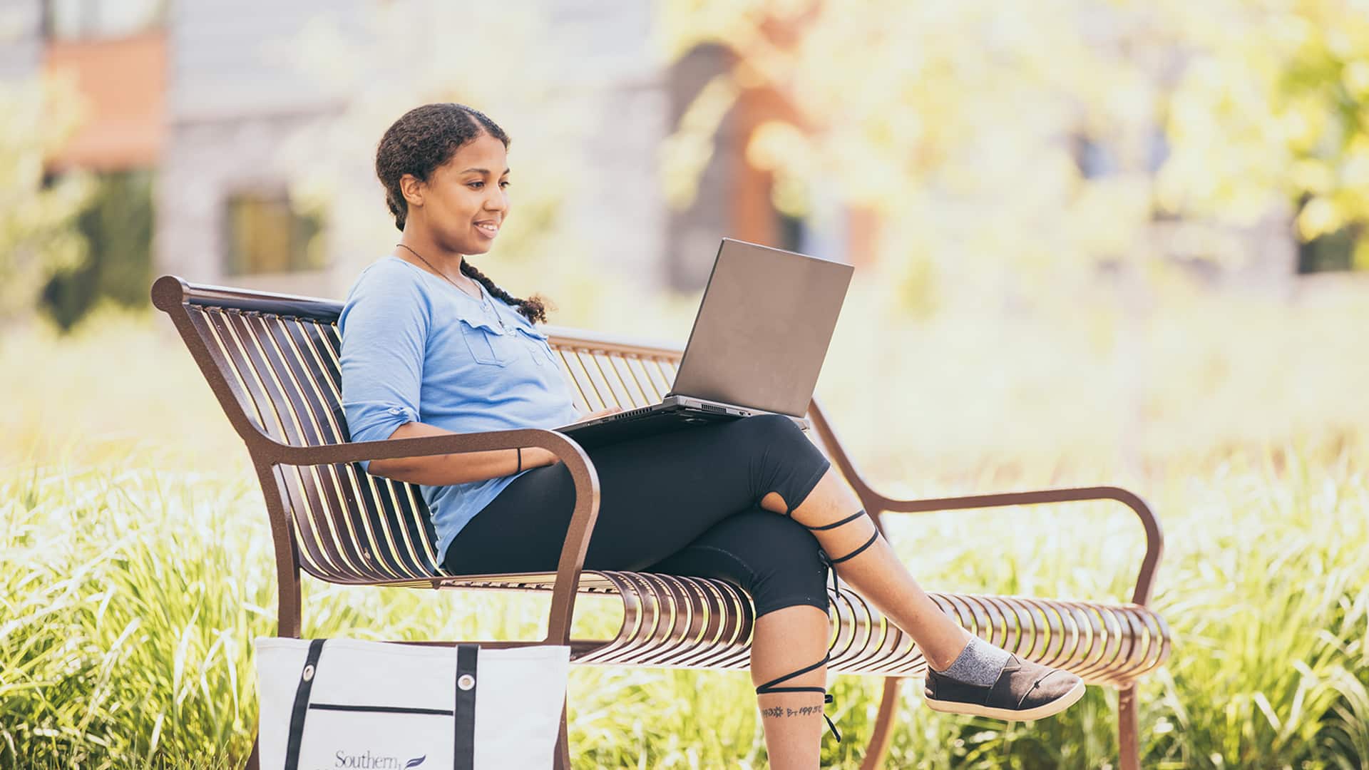 Bethanie Thomas sitting on a park bench working on her laptop with an SNHU shoulder bag on the ground next to her.