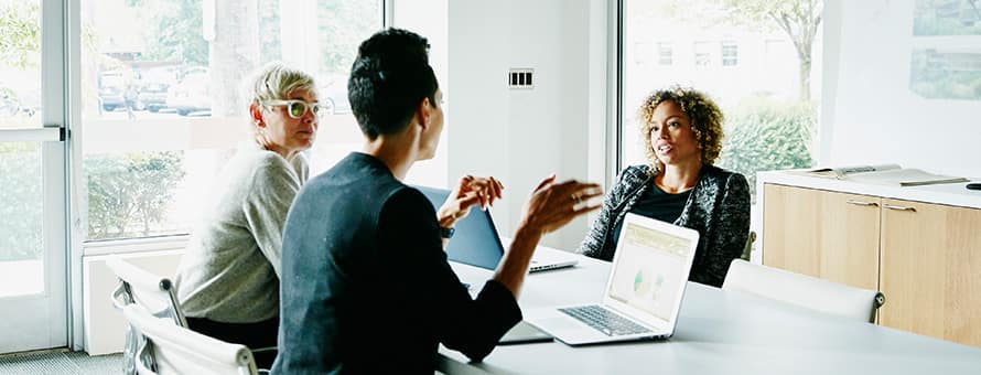 Three coworkers sitting around a table at a meeting with their laptops open for their job they got with their business degree