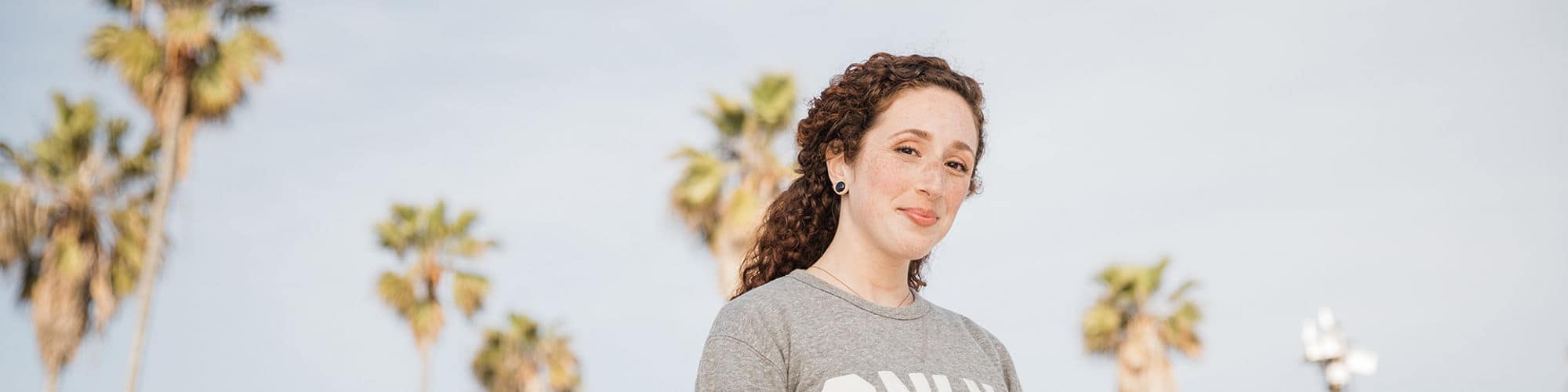 California resident Mariel Embry, who earned her degree in creative writing and English from SNHU, wearing a gray SNHU sweatshirt with tall palm trees behind her.