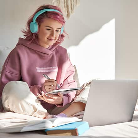 A person wearing a pink sweatshirt and teal headphones, researching on a laptop which graduate program to choose.
