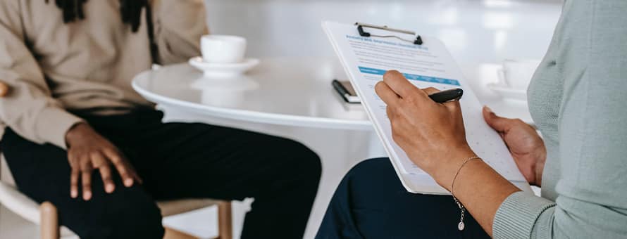 A clinical mental health counselor taking notes on a clipboard while talking to a client.
