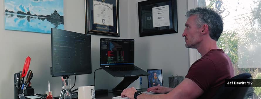 Jef Dewitt '22, who graduated from an online computer science program at SNHU, working on laptop and monitor at desk from home..