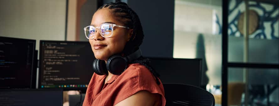 A master's in computer science student with over-ear headphones around her neck, sitting at a desk with code on the monitor.