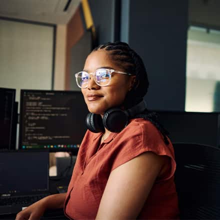 A master's in computer science student with over-ear headphones around her neck, sitting at a desk with code on the monitor.