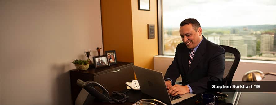 Stephen Burhart, wearing a dark suit, typing on a laptop in an office with a large window behind him overlooking city buildings.