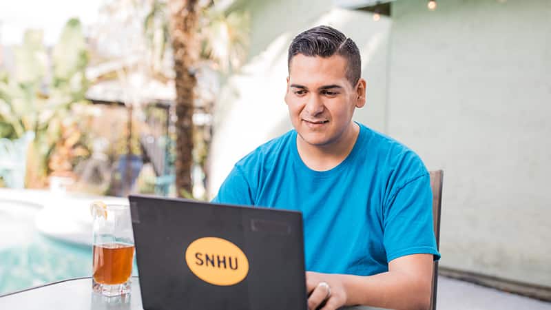Juan Munoz, who earned his degree from SNHU in 2022, sitting at an outdoor table and typing on his laptop.