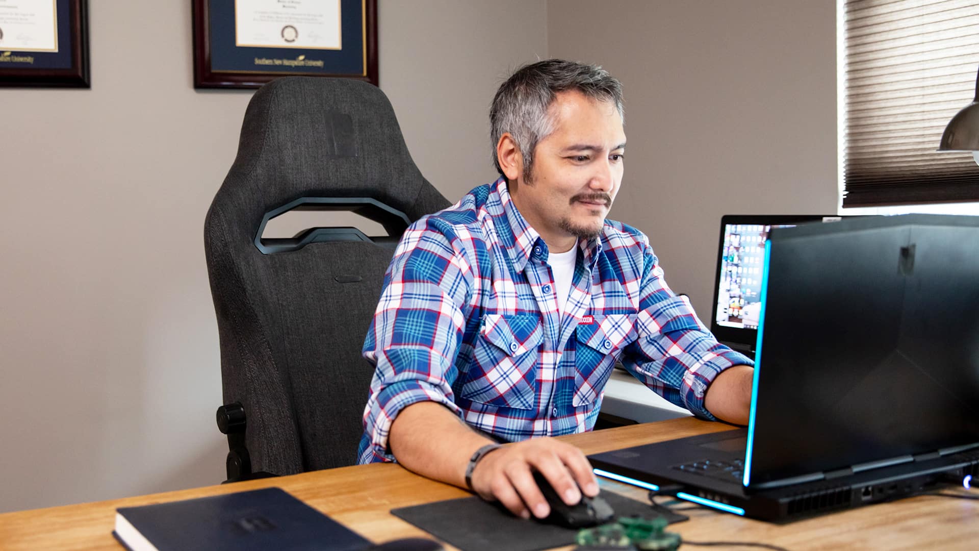 SNHU graduate Derald Wise sitting at a desk and working on a laptop with his framed diploma on the wall behind him.