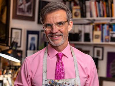 Dale Stephanos, who earned his graphic design degree from SNHU, wearing a pink button down shirt and tie and a paint spattered apron.