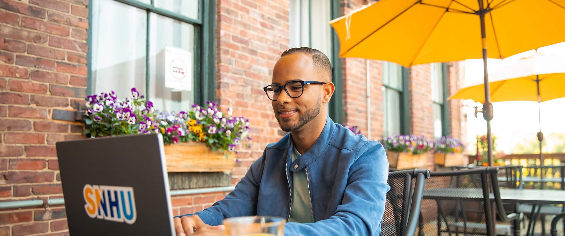 Geremy Laboy Prospere, sitting at an outdoor cafe working on a laptop with an SNHU sticker on the cover.