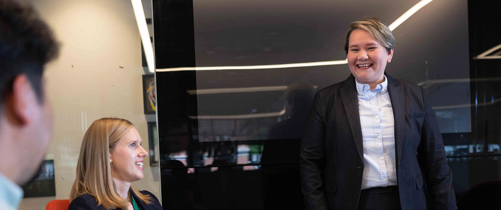  A woman with short hair wearing a dark blazer and speaking to a man and woman in a conference room.